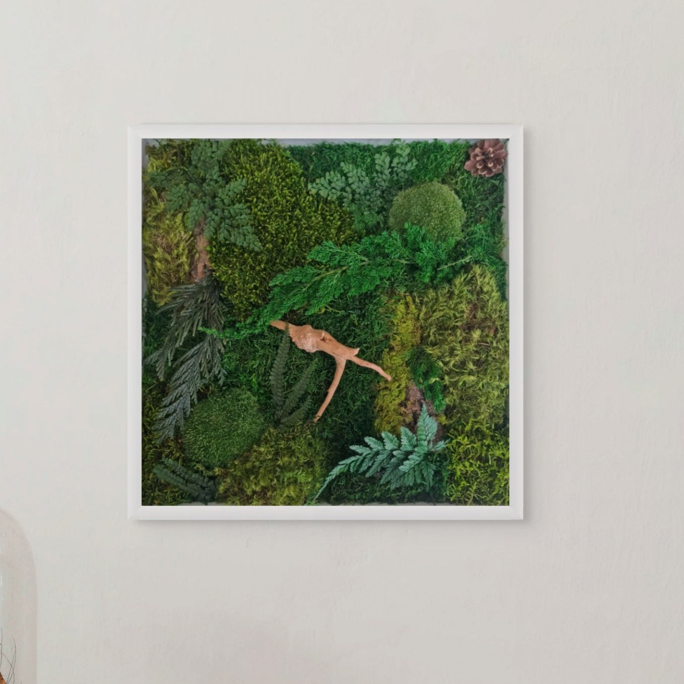 A framed artwork featuring a forest floor scene with moss, ferns, and a figure lying down, set against a white background.
