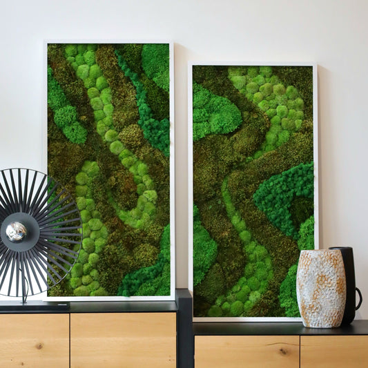 Two framed artworks consisting of preserved moss in various shades of green, displayed on a wooden console table against a white wall, with decorative objects beside the frames.