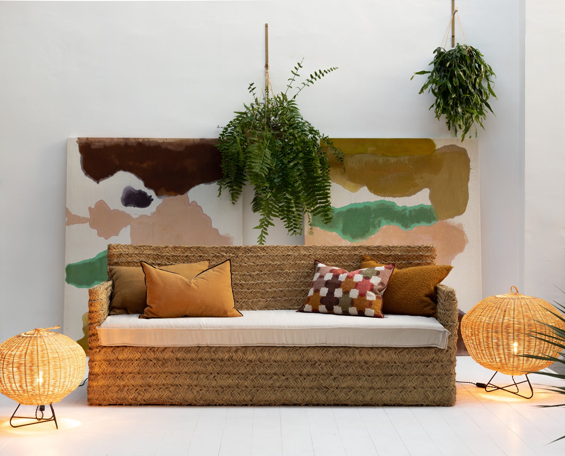 woven sofa  made from natural fibre, esparto, with long white seating cuschion and decorative colorful cushions against a white wall with decorative plants and painting